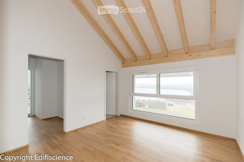 Empty room with wooden flooring, white walls, and large windows offering a view. The ceiling features exposed wooden beams.