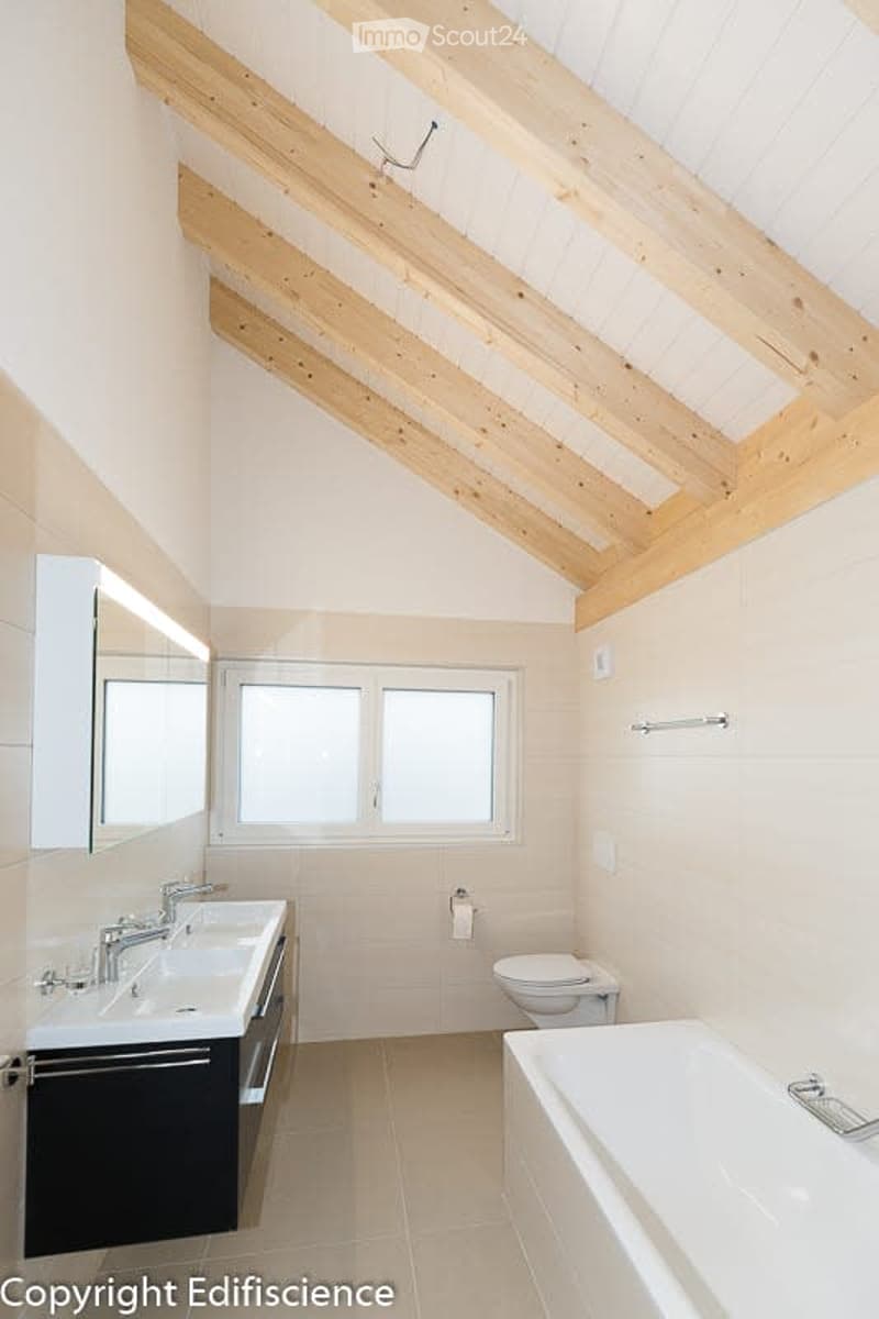 Modern bathroom with exposed wooden beams on the ceiling, white tiles, double sink, bathtub, toilet, and large windows providing natural light.