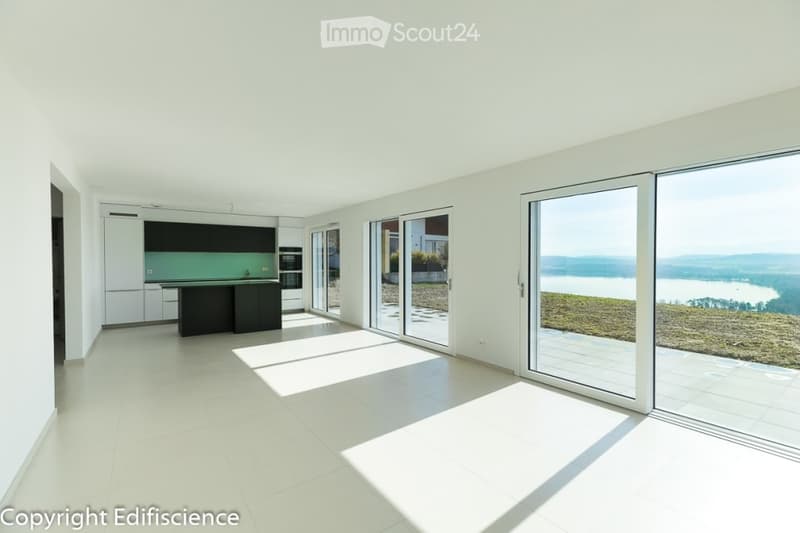 Modern, open-plan kitchen and living space with large windows offering a scenic view, white cabinets, central island, and tiled flooring.