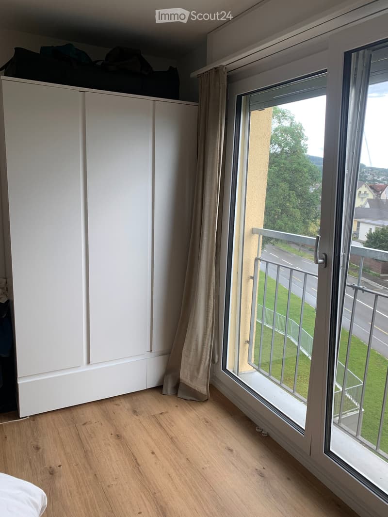 Empty room with wooden flooring, white built-in wardrobe, large window with balcony access, and light-colored curtains.