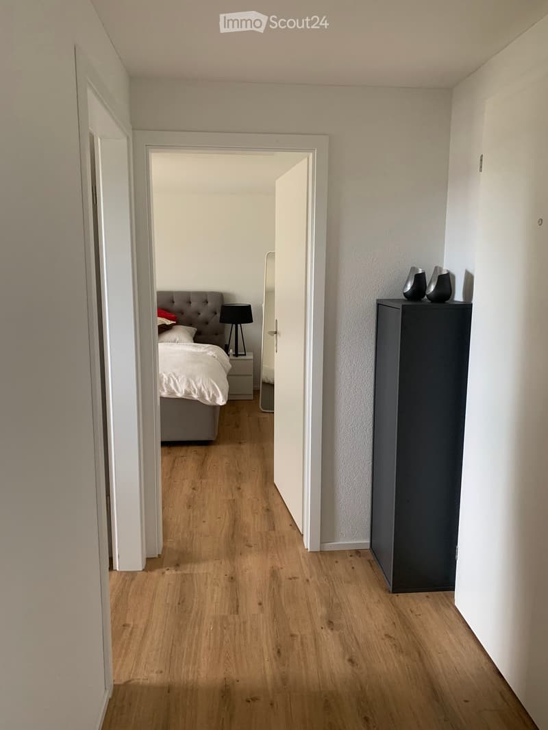 A narrow hallway with light wooden flooring and white walls, leading to a bedroom with a grey upholstered headboard, white bedding, and a bedside table with a lamp. A small black cabinet with decorative vases is visible on the right side.
