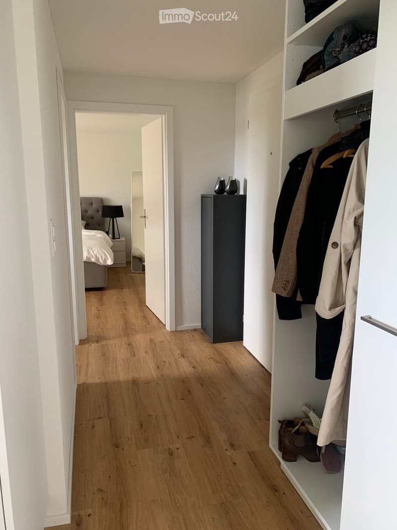A hallway leading to a bedroom with a wooden floor, white walls, and a built-in wardrobe with hanging clothes and shoes. The bedroom features a bed, a lamp, and a small table.