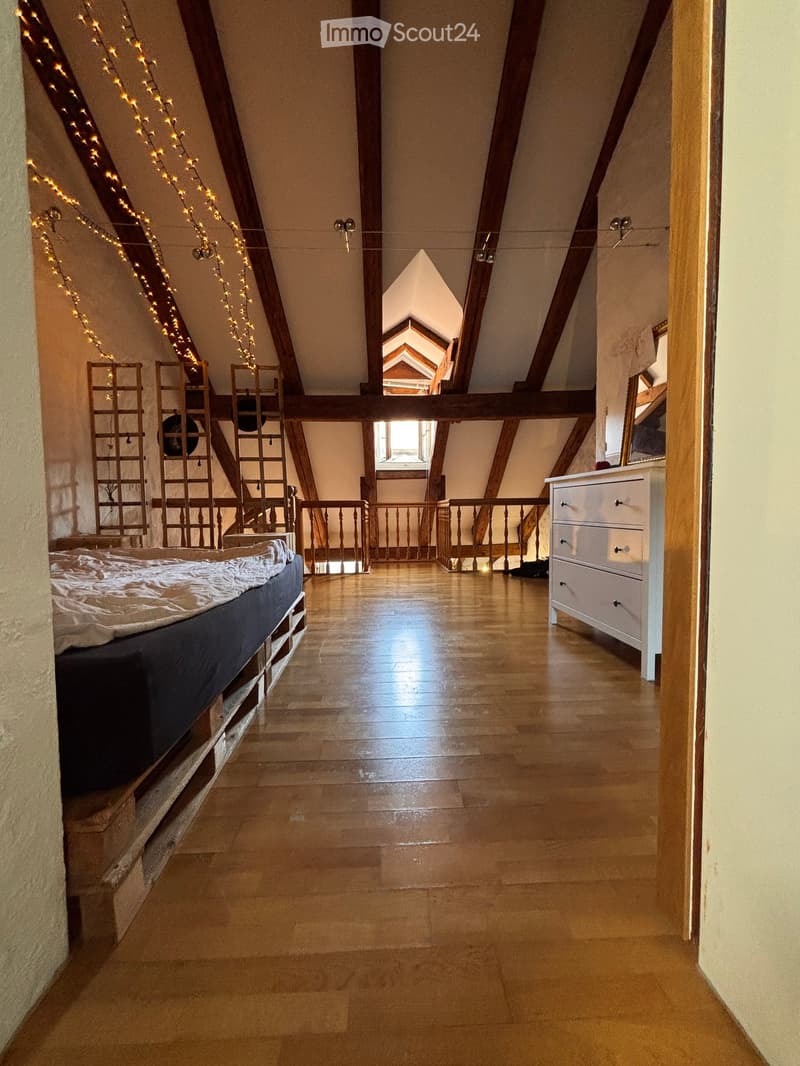 Cozy attic bedroom with wooden beams, exposed wooden floor, and a bed on the left side. Features a small balcony area with wooden railings and string lights.