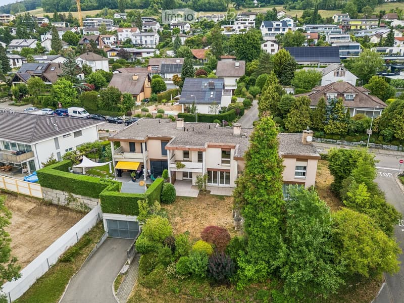 Aerial view of a suburban neighborhood with multiple houses, gardens, and parked cars. Houses feature varied architectural styles, some with solar panels on the roofs, surrounded by greenery and trees.
