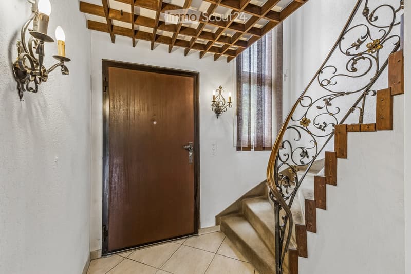 Elegant hallway with a wooden door, ornate wall-mounted candle holders, and a staircase with a decorative wrought-iron railing and wooden steps.