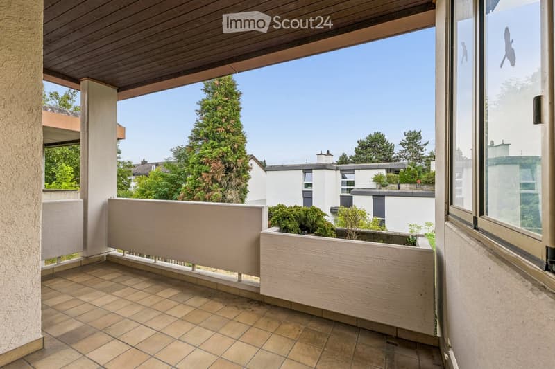 Balcony with tiled floor, wooden planter, and wooden railing, overlooking a green area with trees and neighboring houses.