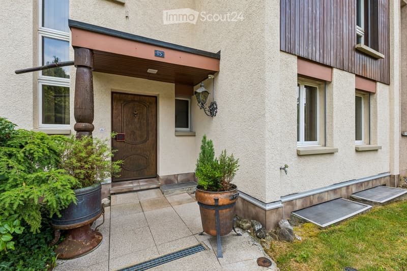 Front entrance of a two-story building with a wooden door, decorative columns, potted plants, and a small garden area.