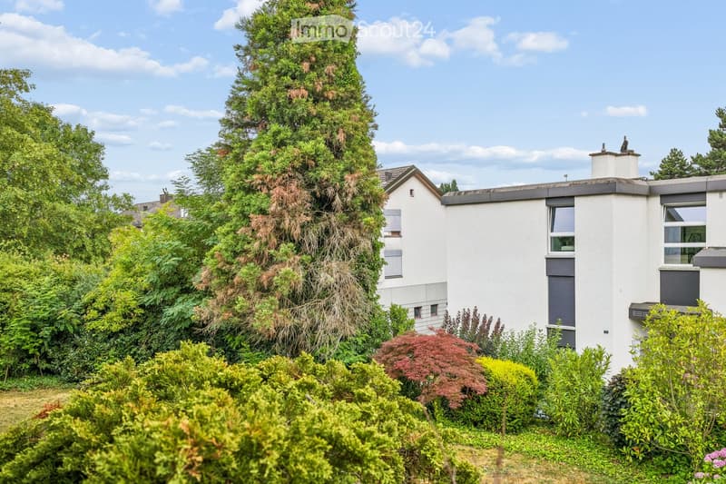 A modern two-story house with white walls, grey accents, and a chimney, surrounded by lush greenery and trees.