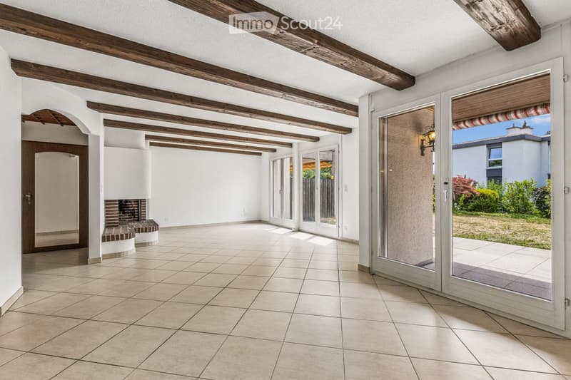 Bright, open space with exposed wooden beams, tiled floor, and large glass doors leading to an outdoor area. Features a built-in brick fireplace and white walls.