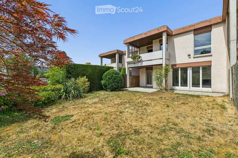 A two-story house with a modern design, featuring large windows, a balcony, and a well-maintained garden with shrubs and trees.