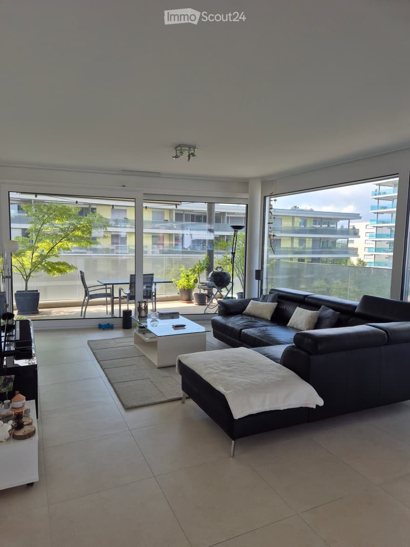 Modern living room with large windows and balcony access, featuring a black leather sofa, coffee table, ottoman, side table, and potted plants.