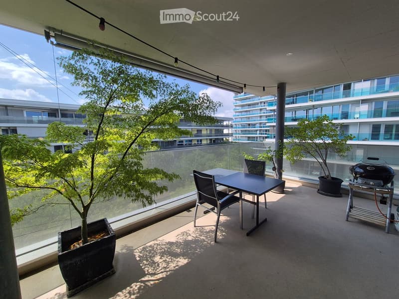 Modern balcony with a dining table, chairs, grill, and potted plants, overlooking a cityscape with contemporary buildings.