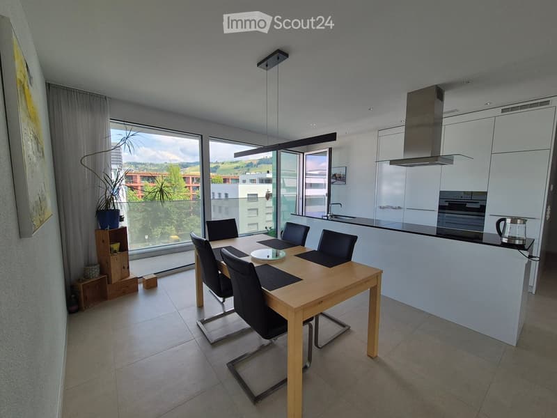 Modern open-plan kitchen and dining area with a wooden dining table, black chairs, white kitchen cabinets, and a large window overlooking a green landscape.