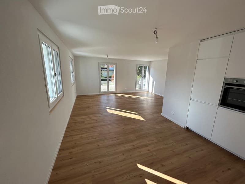 Empty room with wooden flooring, white walls, and large windows allowing natural light to enter. The room features built-in white cabinetry on one side and a partially visible oven.
