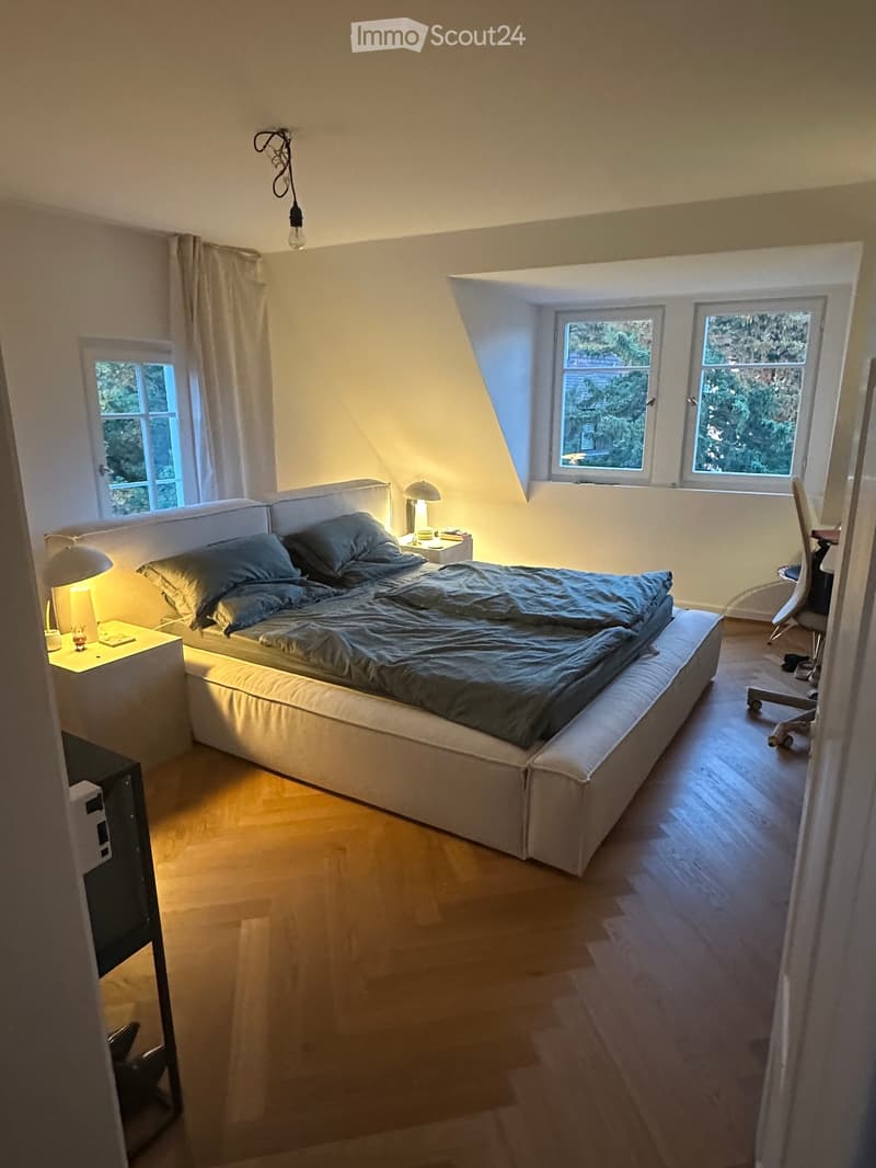 Cozy bedroom with a large bed, nightstands, lamps, and a chair near the window. The room has wooden flooring and large windows with a view of greenery.