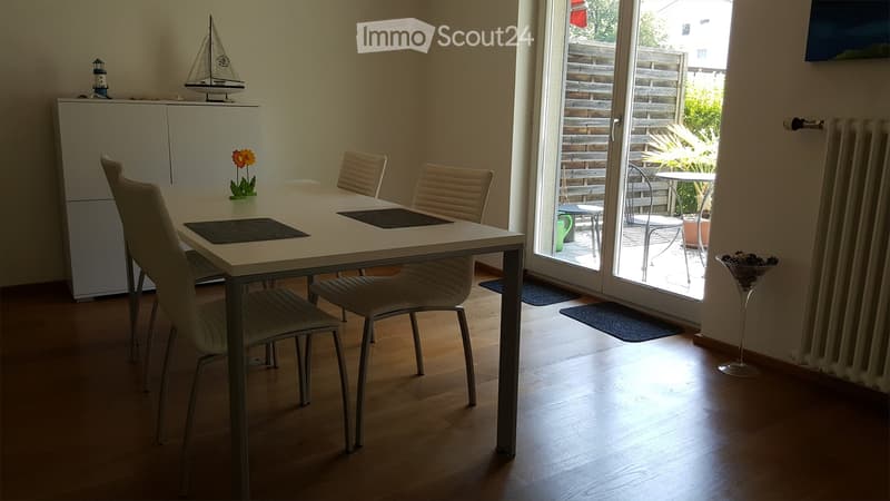 Bright dining room with a white dining table, four beige chairs, a white cabinet, a small flower vase, and a model sailboat on top of the cabinet. A large sliding glass door leads to a patio with outdoor furniture.