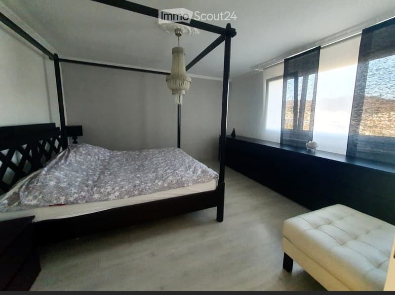 A modern bedroom with a four-poster bed, a chandelier, a nightstand, and a white leather stool.