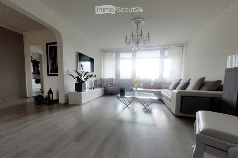 Modern living room with light-colored walls, wooden flooring, a large sectional sofa, coffee table, armchair, chandelier, television, and decorative plants.