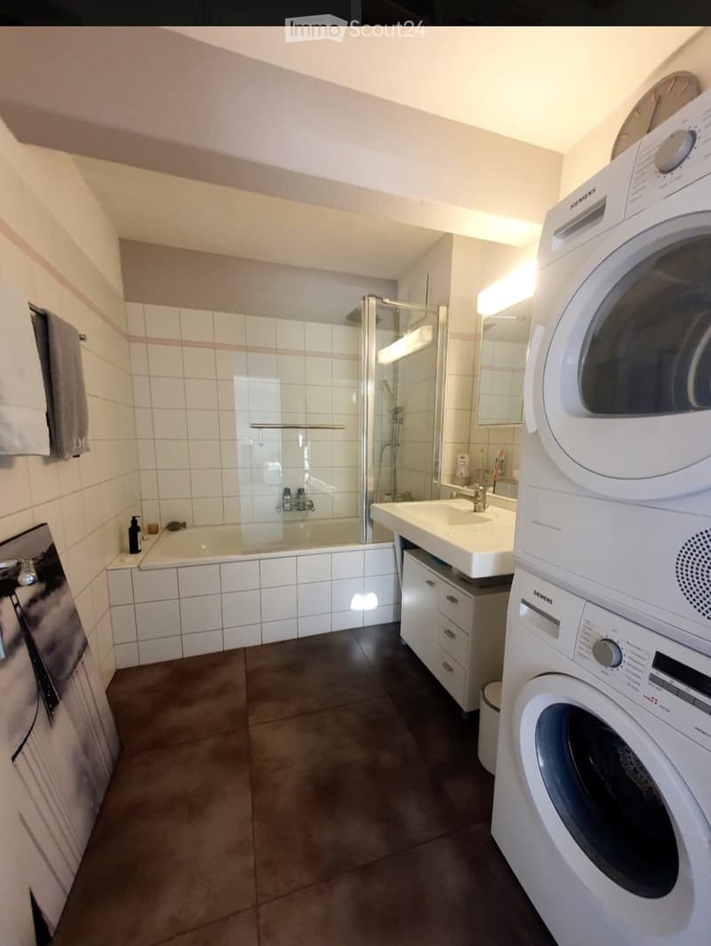 Modern bathroom with a bathtub, shower stall, sink, mirror, and a washing machine. The space features white tiled walls, a dark tiled floor, and a towel hanging on the wall.