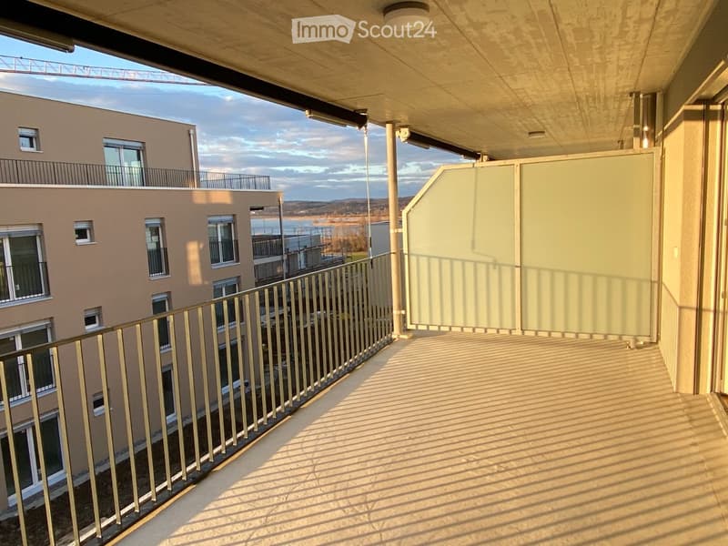 Balcony with metal railing, overlooking a modern building complex and a body of water in the distance.