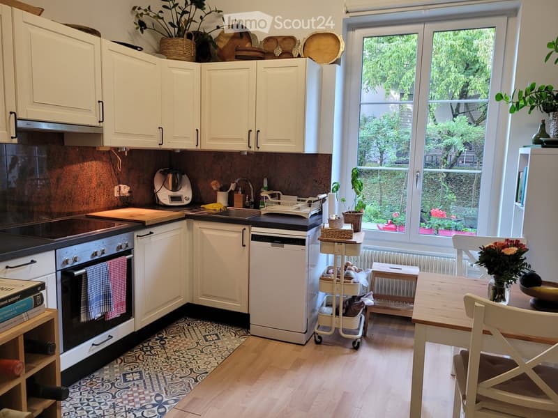 Modern kitchen with white cabinets, stainless steel appliances, and a large window overlooking a garden. Includes an oven, dishwasher, sink, cutting board, blender, cart, dining table, chairs, and decorative items.