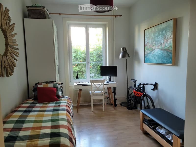 Cozy bedroom with a single bed, desk, chair, bicycle, and a stool. Features a large window with a view of greenery, a hanging lamp, and a picture frame on the wall.