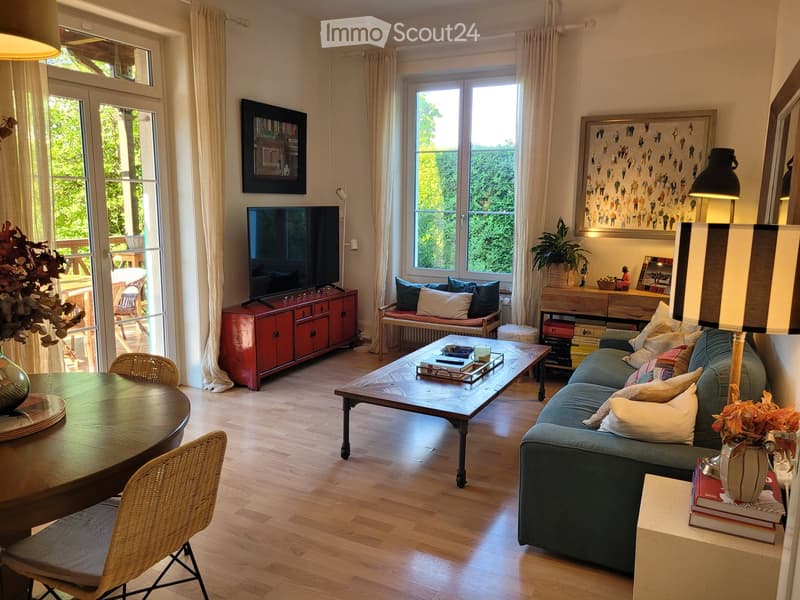 Bright living room with large windows and a view of greenery, featuring a blue sofa, wooden coffee table, dining table with chairs, TV on a red cabinet, and decorative plants.