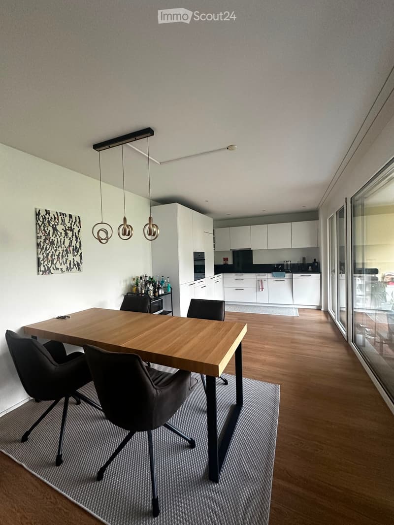 Modern kitchen and dining area with a wooden dining table, black chairs, pendant lights, white cabinets, and a small bar area with bottles and cups.
