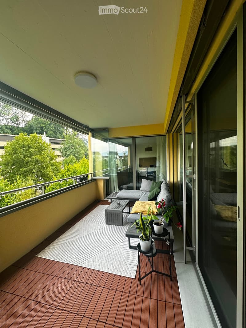 Balcony with wooden decking, white rug, sofa, small table, potted plants, and a view of greenery.
