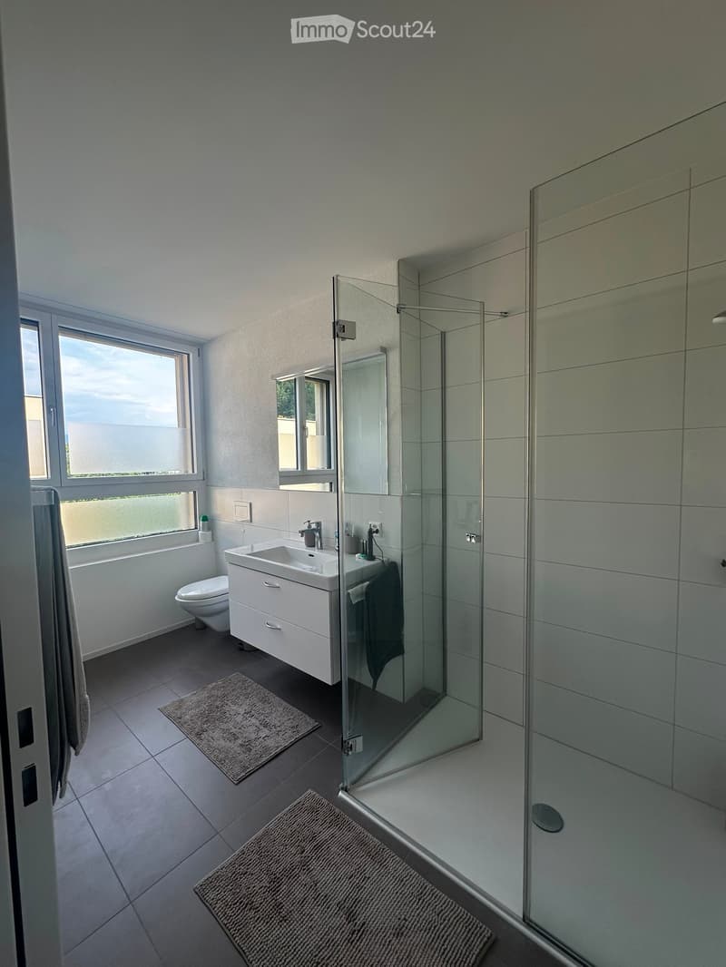 Modern bathroom with white tiles, a glass-enclosed shower, a white sink with a chrome faucet, a toilet, and two towels on the floor.