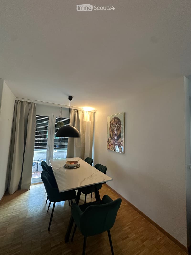 Dining room with a white marble dining table, four green chairs, a pendant light, and a large picture frame on the wall. The room has wooden flooring and large windows with curtains.