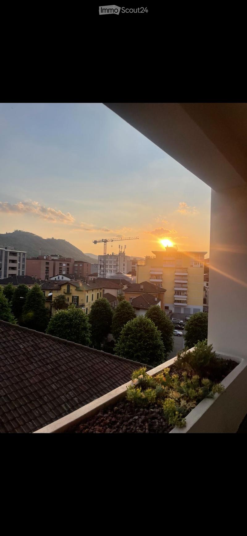 Balcony with small garden bed, overlooking a cityscape at sunset with buildings, trees, and a distant mountain range.