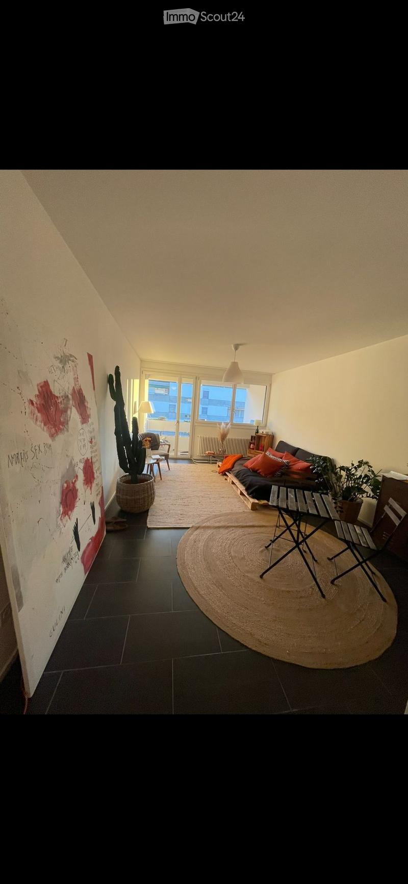A modern living room with a dark tiled floor, a large round jute rug, a sofa with colorful cushions, a potted cactus, a small table, and a hanging lamp.