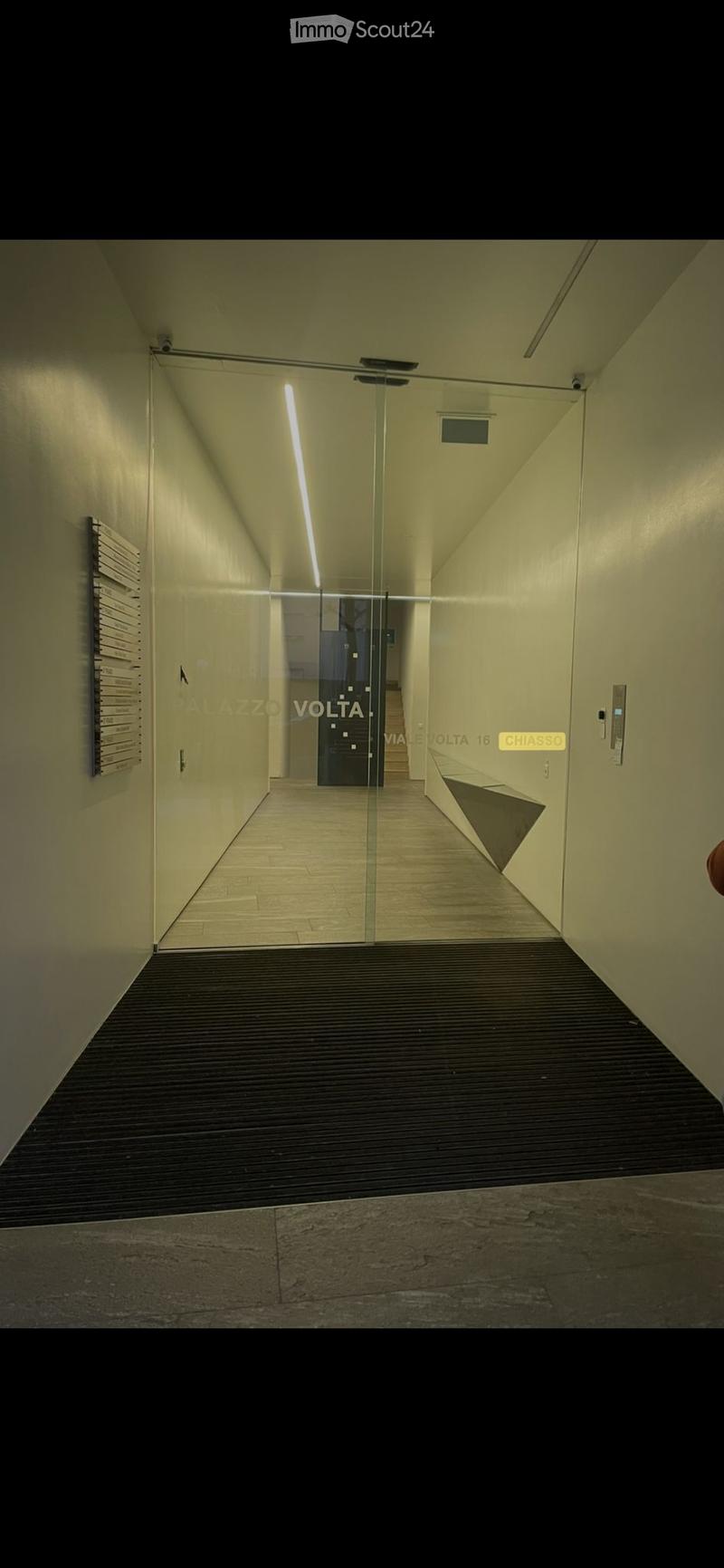 Modern hallway with glass partition, light-colored walls, and tiled flooring. Features a sleek design with a visible elevator area in the background.