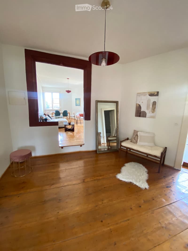 Empty room with wooden flooring, white walls, a large mirror, a hanging lamp, and a small bench with cushions visible in the background.