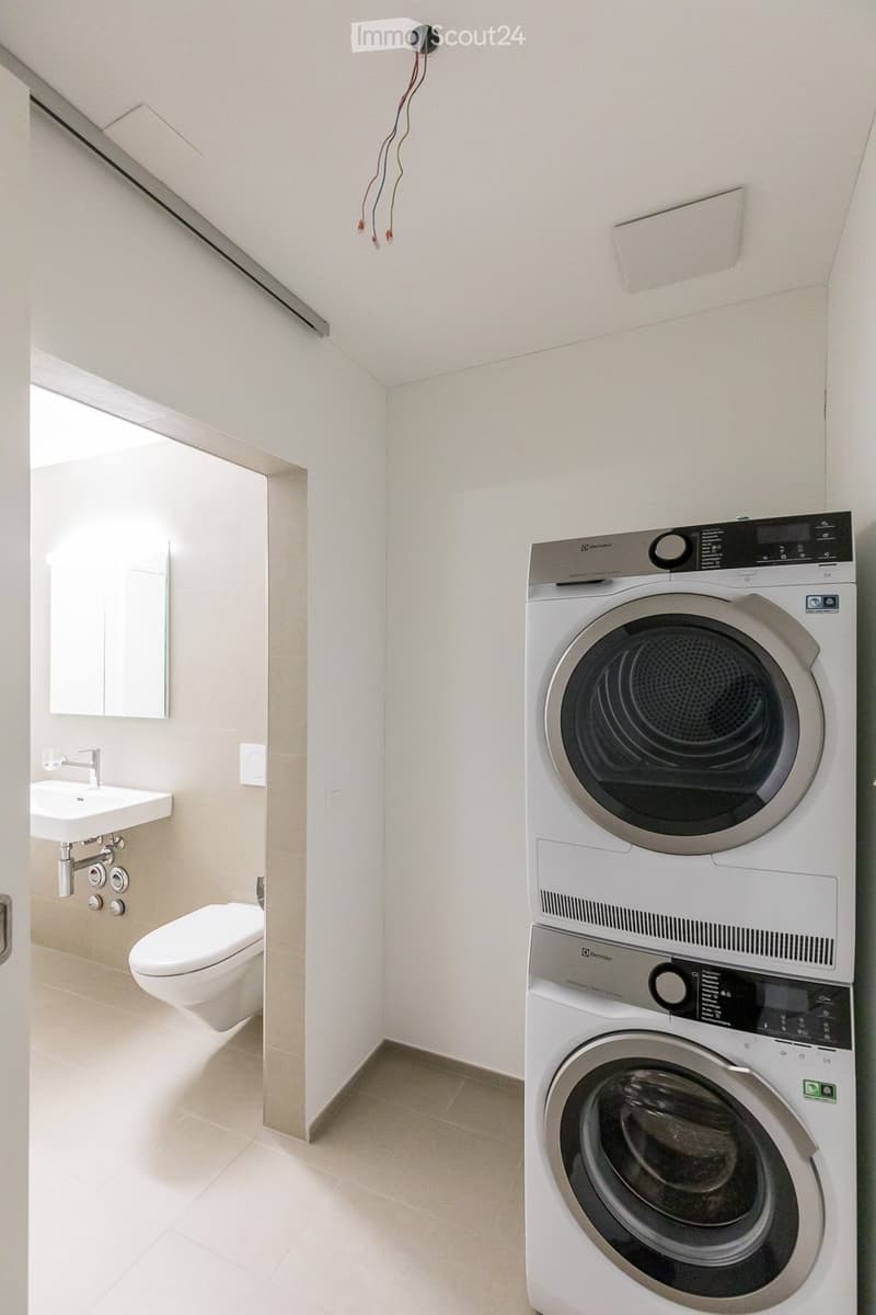 Modern bathroom with a washing machine and dryer unit, a sink with a faucet, a toilet, and a mirror. The space is clean and well-lit with light-colored tiles and walls.