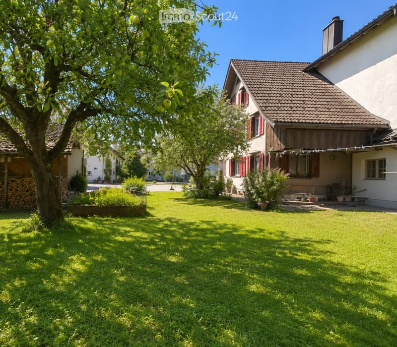 Lush green lawn with a large tree, a wooden house with red shutters, and a stack of firewood on the left side.