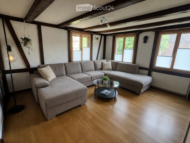 Modern living room with wooden flooring, large sectional sofa, glass coffee table, floor lamp, and potted plant. Large windows provide natural light.