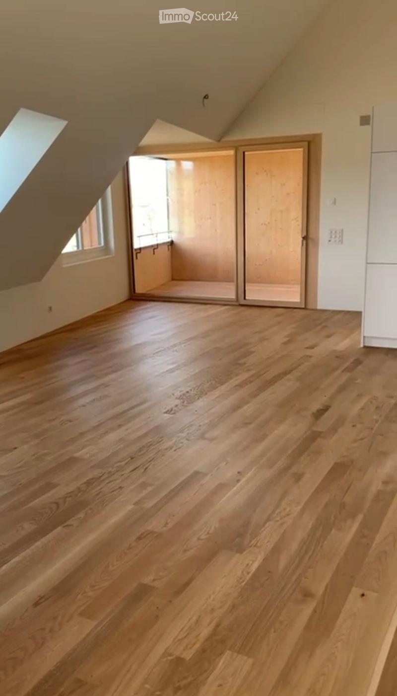 Empty attic room with wooden flooring, slanted ceilings, and large windows, featuring a sliding door leading to another space.