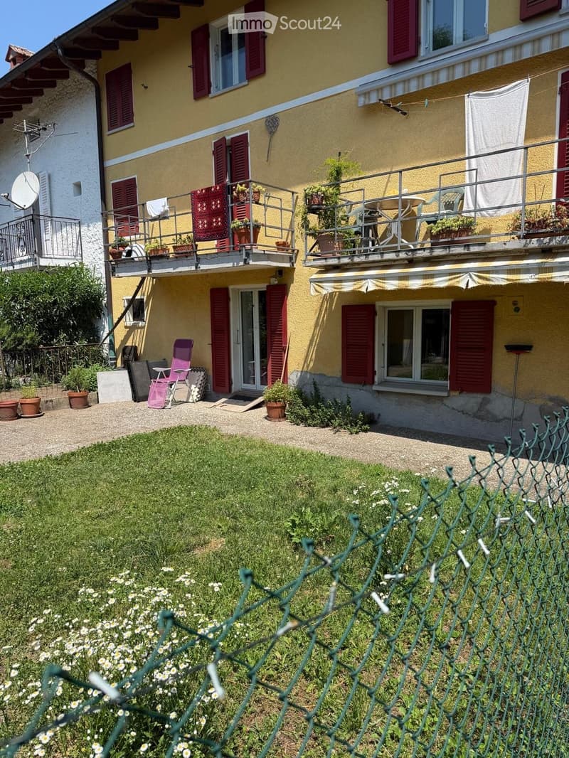Two-story yellow house with red shutters, balconies, outdoor seating, and garden area.