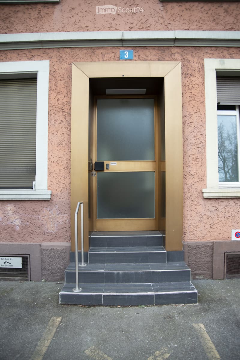 Entrance to a building with a raised ground floor, featuring a set of stairs leading to a glass door framed in wood, flanked by two windows with white frames.