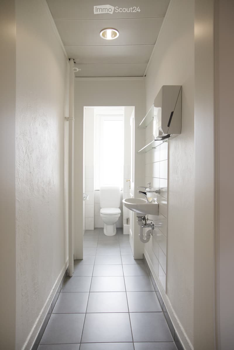 A clean, modern bathroom with white tiled walls and floor, a toilet, a sink with a faucet, and a mirror above it. The space is well-lit with a ceiling light fixture.