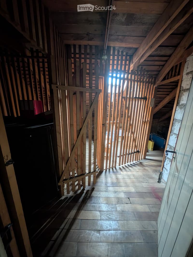 An unfinished attic corridor with exposed wooden beams and a staircase leading upwards, featuring a wooden railing and tiled floor.