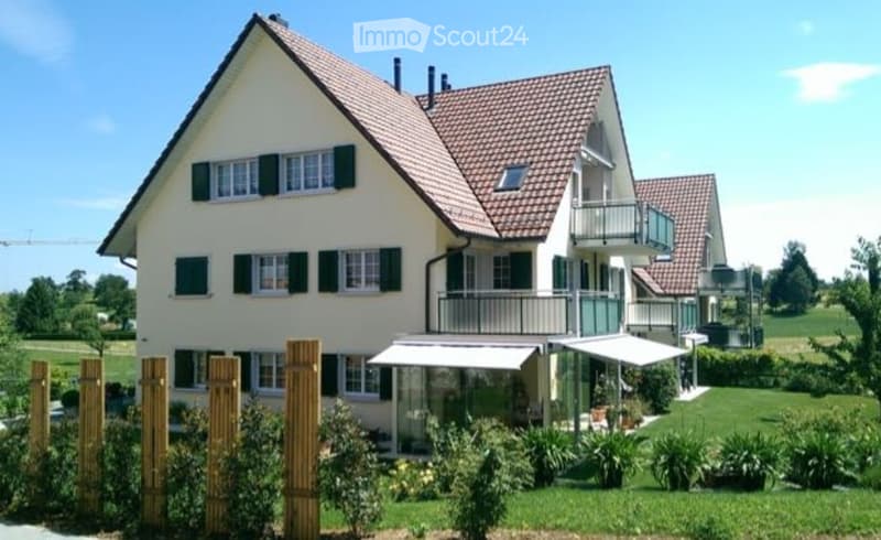 Two-story house with a red-tiled roof, white walls, multiple windows with shutters, and a balcony on the upper level. The house is surrounded by a well-maintained garden with greenery and a covered patio area.