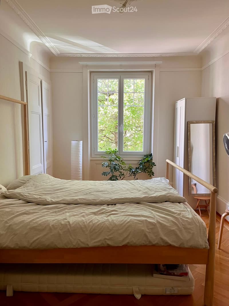 Cozy bedroom with a double bed, wooden floor, large window with a view of greenery, and a small plant on the windowsill.