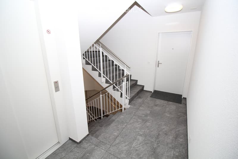 Modern hallway with grey tiled floor, white walls, staircase with metal railing, and a closed door.