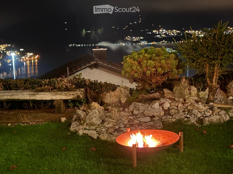 Nighttime garden with a fire pit, stone rocks, greenery, and a cityscape view in the background.