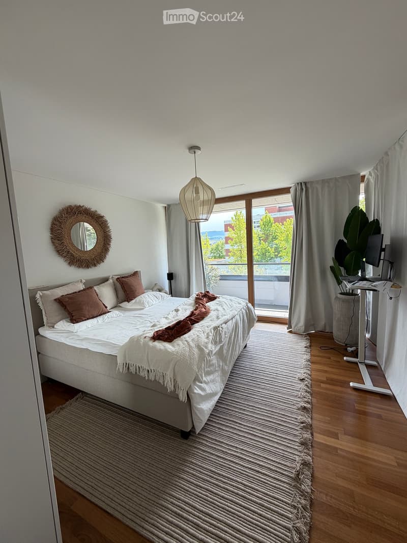 Modern bedroom with a large bed, white bedding, throw blanket, pillows, a round mirror, a hanging pendant light, a desk with a laptop, and a plant near the window offering a view.
