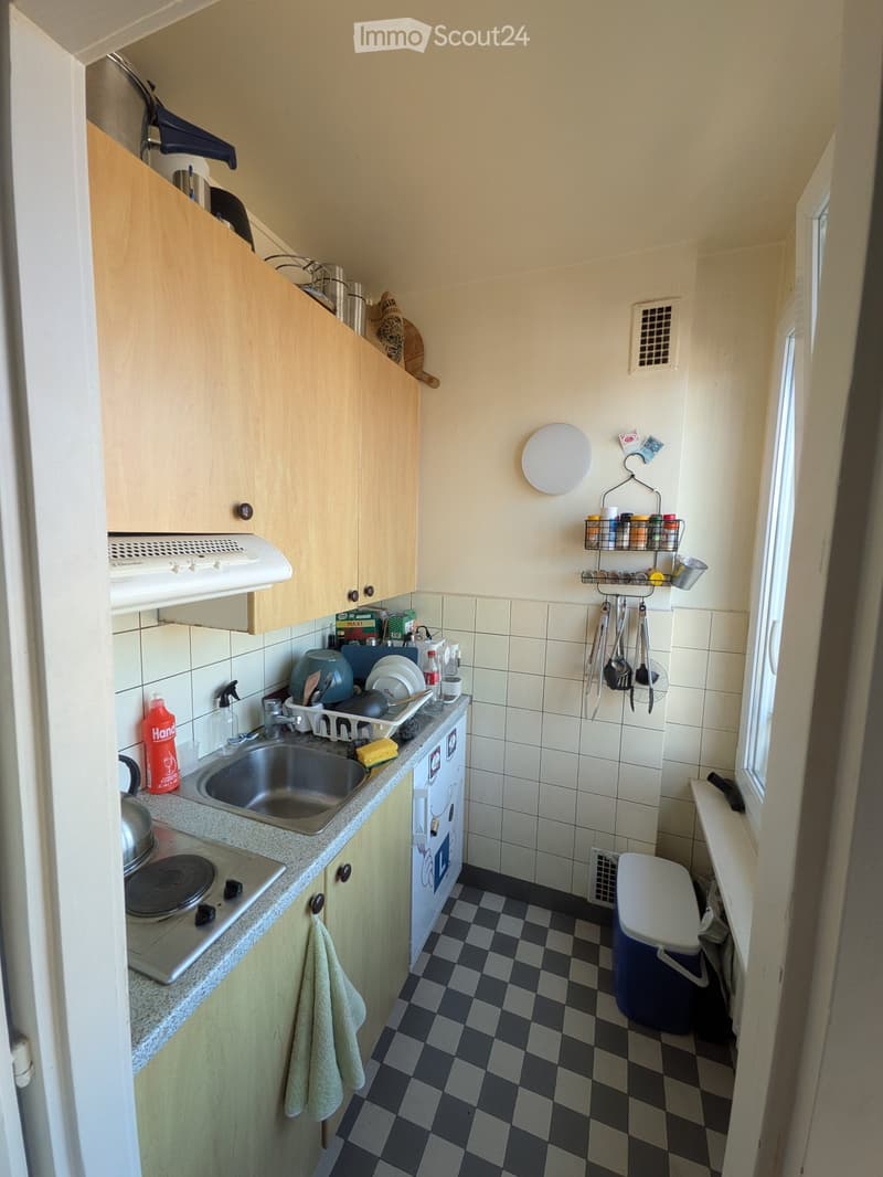 Compact kitchen with light wooden cabinets, a sink, stove, and various kitchen items on the counter and shelves.