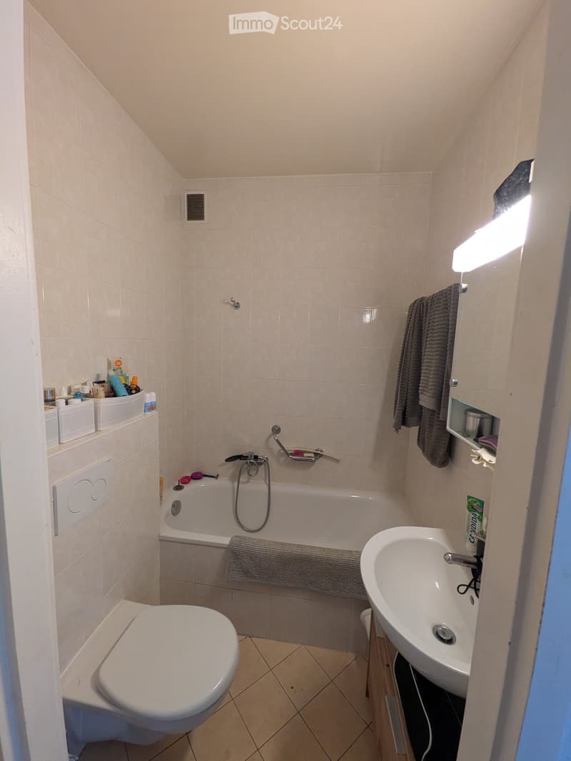 A compact bathroom with a bathtub, toilet, sink, and towel. The walls are tiled in light beige, and the floor is tiled in a similar color. A towel hangs on the side of the bathtub, and a small shelf holds various toiletries.